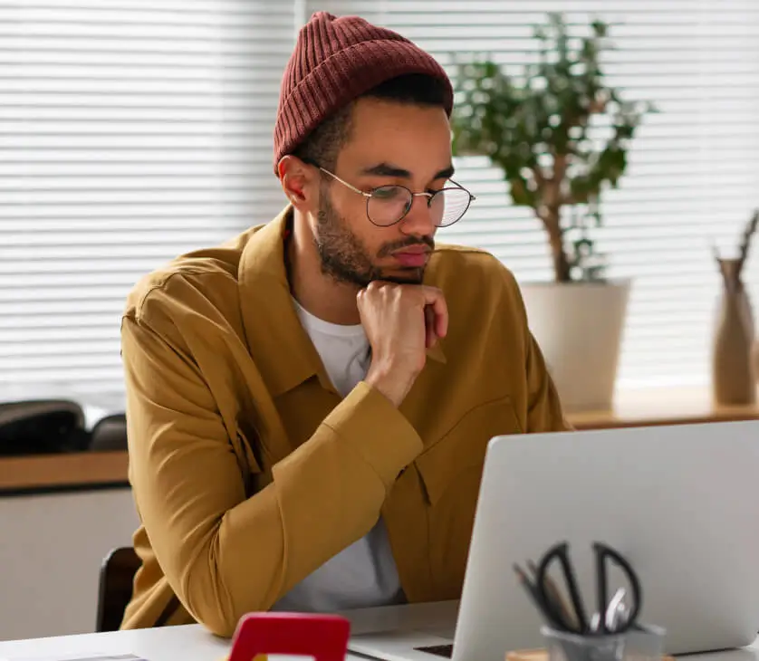 A person wearing glasses, a red beanie, and a brown shirt sits at a desk, looking thoughtfully at a laptop with the Charleston Website open. Office supplies and a potted plant add to the cozy background.