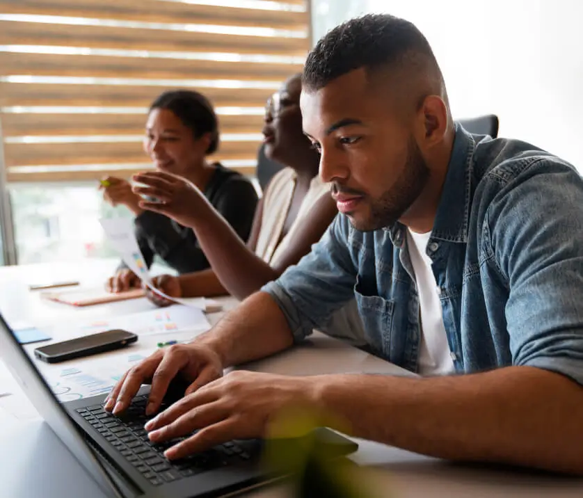 Three people sit at a table working together. A man in the foreground types on a laptop, possibly updating a Charleston Website, while two women in the background smile and look at papers. They appear to be collaborating in a bright, modern office.